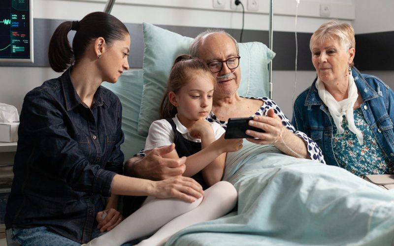 Grandfather browsing on internet with little granddaughter using modern smartphone in hospital ward during clinical therapy. Sick elderly senior man patient resting in bed, healthcare support