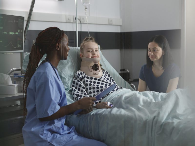 Unwell suffering kid recovering in pediatric hospital ward while resting in patient bed. Nurse examining injured girl with fractured neck wearing cervical collar because of car accident.